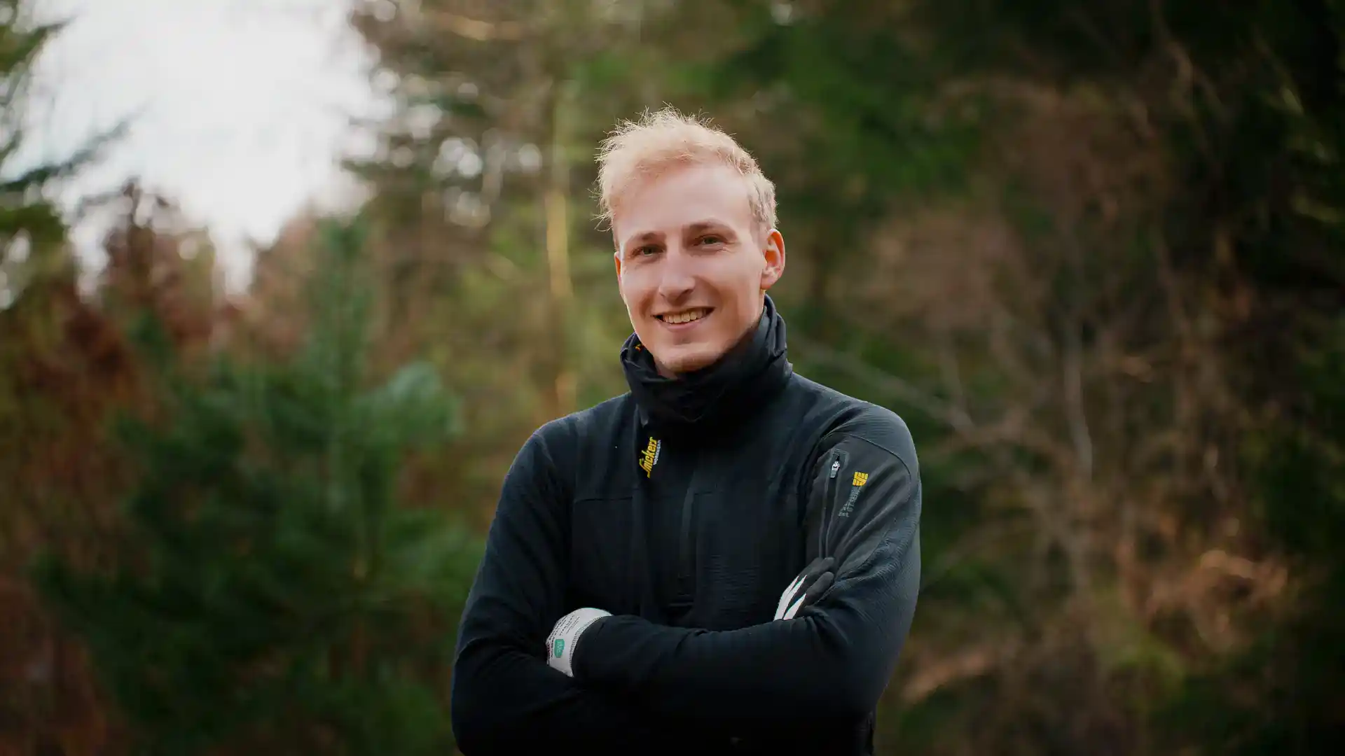 Billedet viser en glad mand i skovarbejde, klædt i sort jakke, med grønne træer i baggrunden.