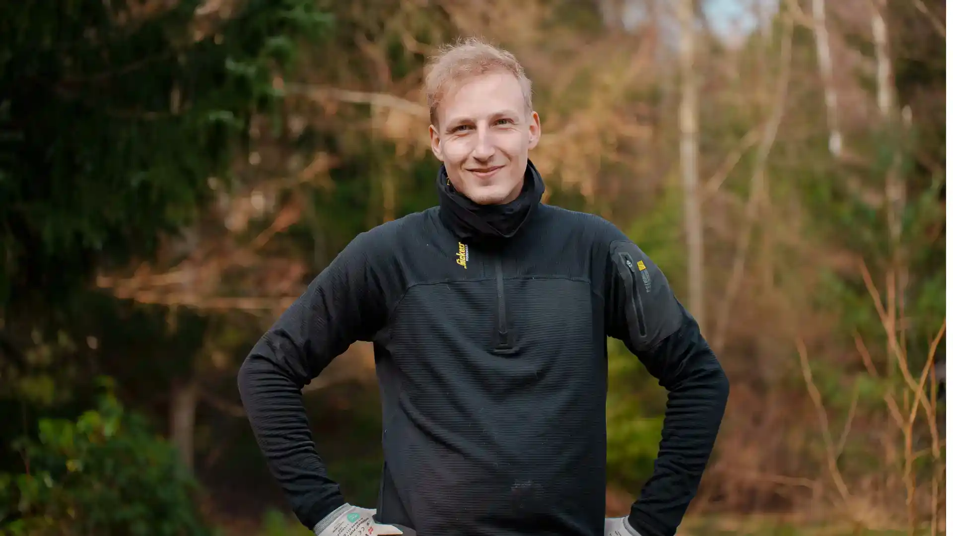 Mands person i outdoor tøj står i skov, smilende, klar til arbejde.