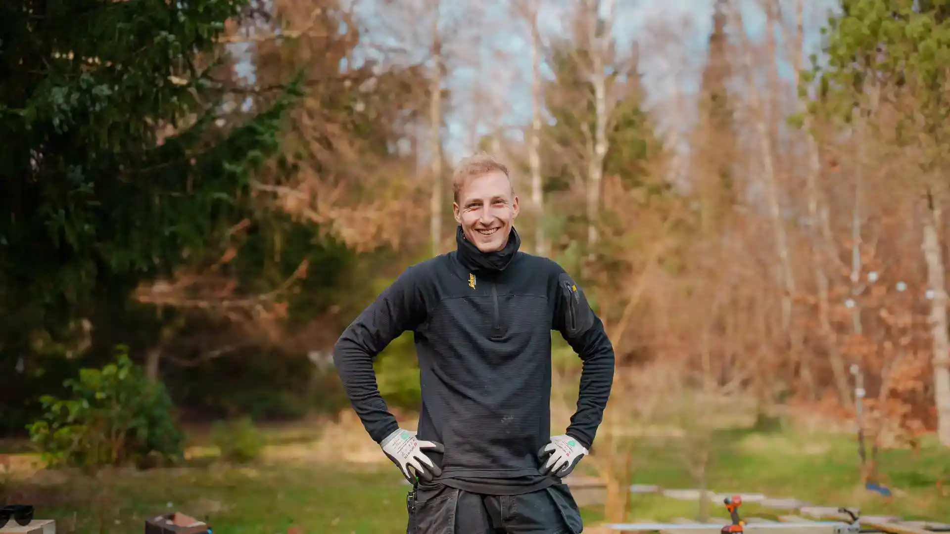 Mand i træarbejdstøj står i skov med træ og værktøj.