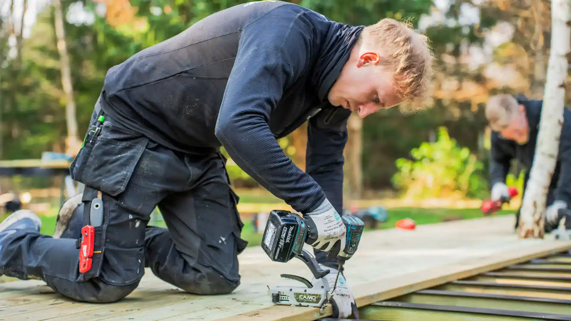 Mand arbejder med sav på træterrasse i udendørs miljø.